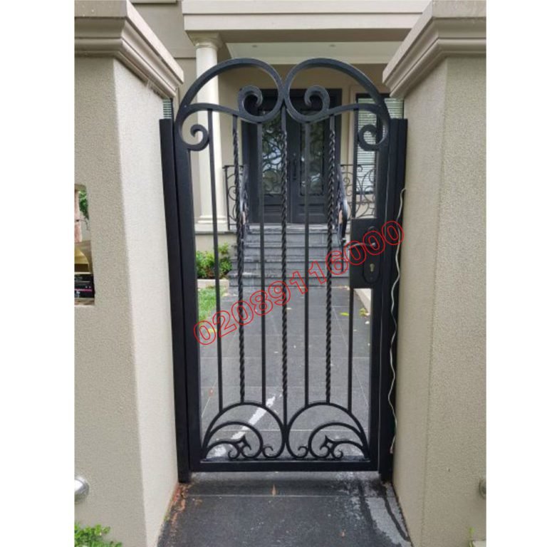 Black arched metal garden gate with scroll details fitted between stone pillars on a side path