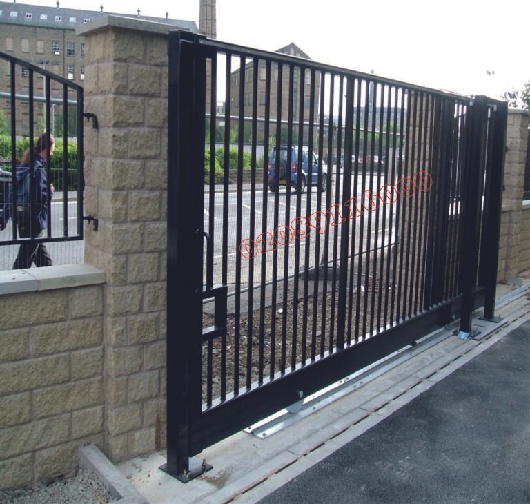 Black metal sliding driveway gate with vertical bars on a rolling track between stone pillars