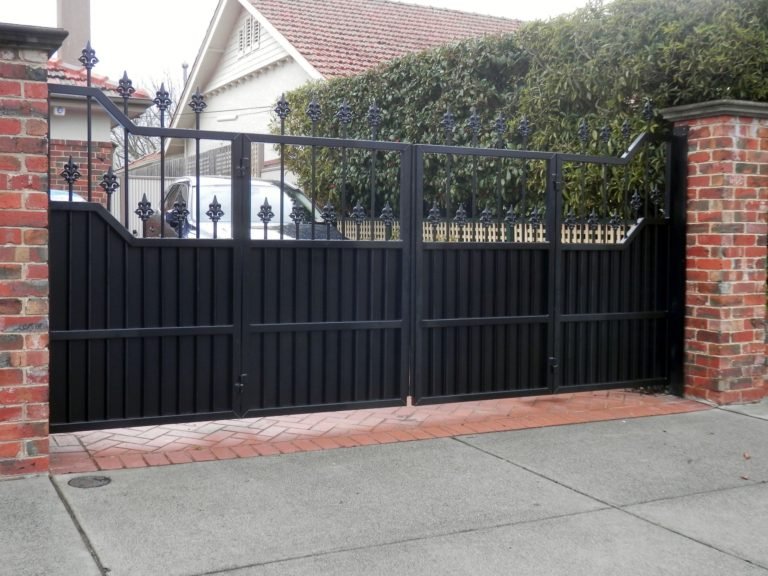Black bifold driveway gate with solid slats and fleur-de-lis top between brick pillars