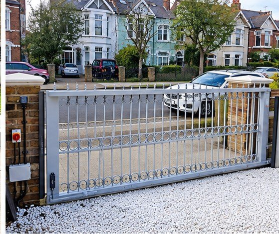 Home 27 Light grey metal driveway gate with vertical bars, circular detailing and pointed finials between brick pillars.
